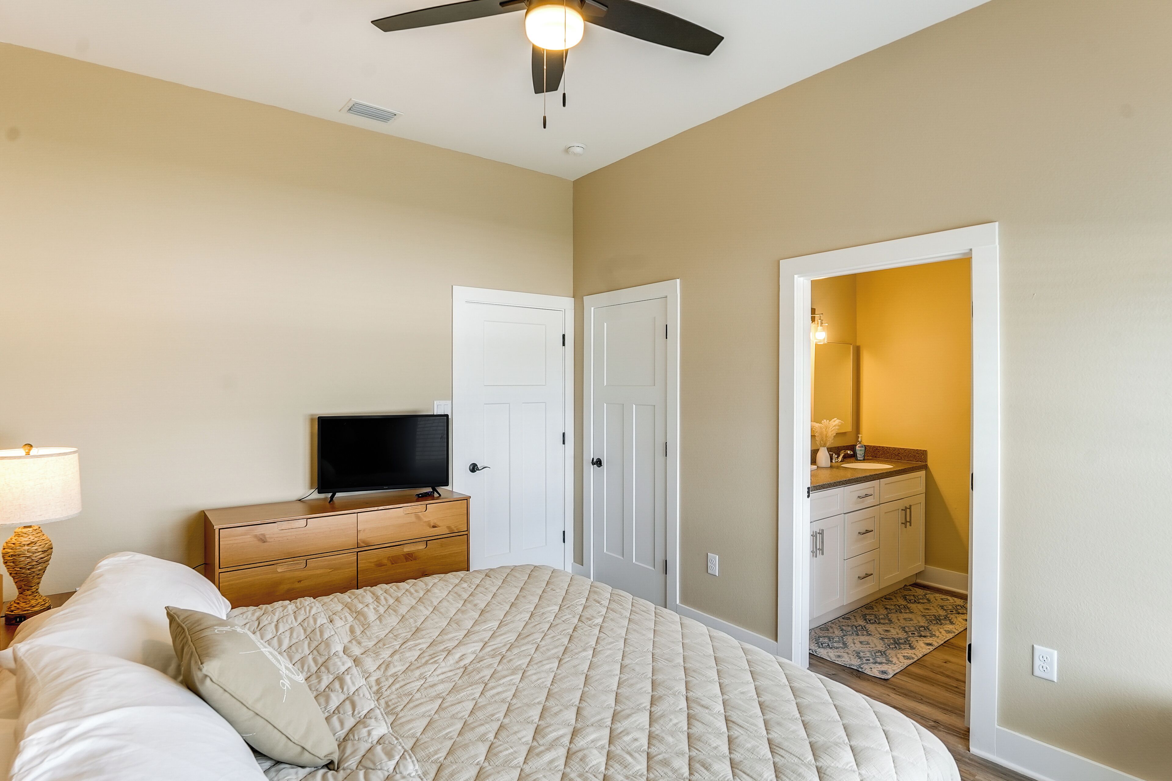 A bedroom with a bed, a TV, a wooden dresser, and a bathroom visible through an open door.