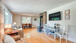 A spacious living room with a fireplace, wooden flooring, a dining area with chairs, and a TV mounted on the wall. - Becket Home w/ 3-season Porch: Walk to Lake Access (Becket)