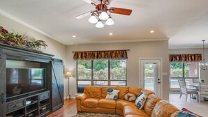 A living room with a sectional sofa, a flat-screen TV, a ceiling fan, and a large window with a view of the outdoors. - Sunny Stephenville Home w/ Pool: 4 Mi to Downtown (Stephenville)