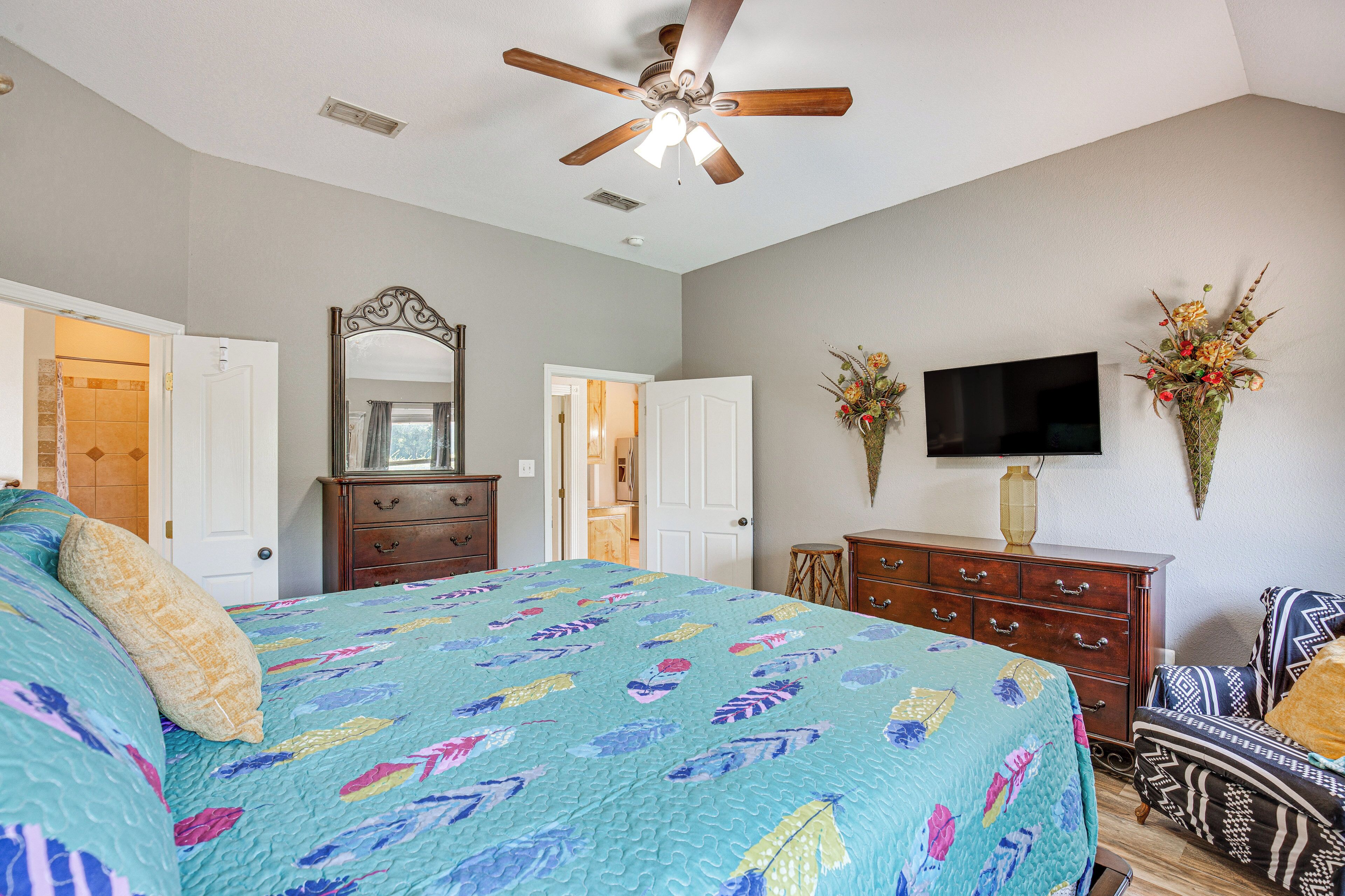 A bedroom with a bed, a ceiling fan, a dresser, a TV, and two flower arrangements.