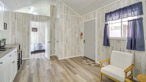 A room with a wooden floor, white walls, a white sofa, and a window with blue curtains. - Picayune Tiny Home Rental ~ 11 Mi to Space Center! (Picayune)