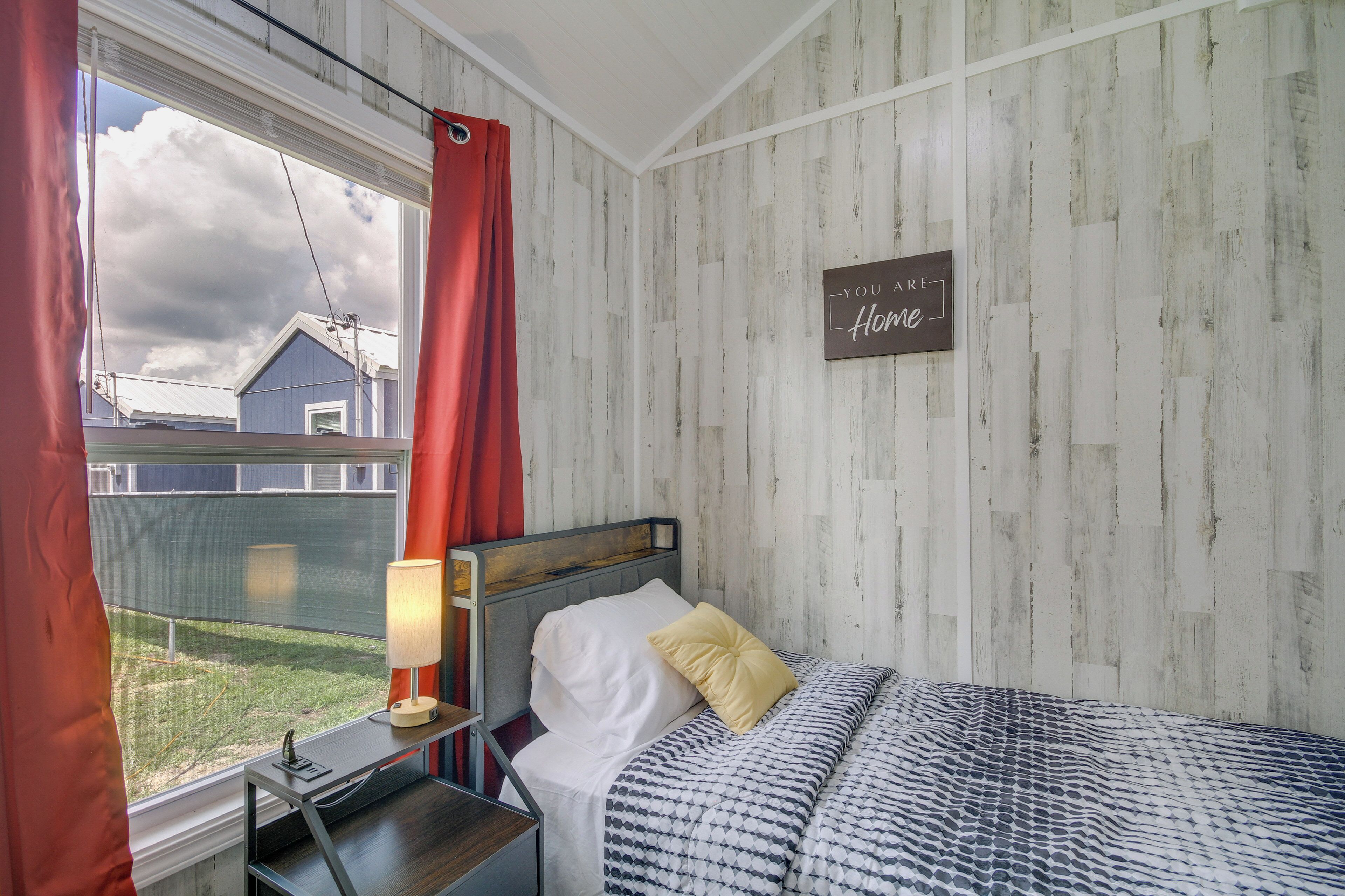 A bedroom with a bed, a nightstand, a lamp, a window with curtains, and a wall with a sign saying "You are home."