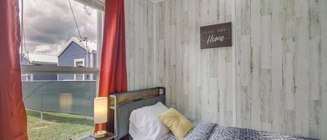 A bedroom with a bed, a nightstand, a lamp, a window with curtains, and a wall with a sign saying "You are home."