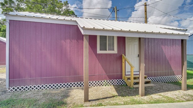 A small, single-story building with a white roof and a purple exterior. It features a front door with a small window above it and a wooden staircase leading up to the entrance. - Picayune Tiny Home Rental ~ 11 Mi to Space Center! (Picayune)
