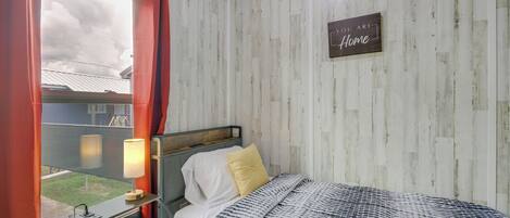 Een slaapkamer met een bed, een nachtkastje met een lamp, een raam met gordijnen, en een bord met de tekst 'You are Home' aan de muur.