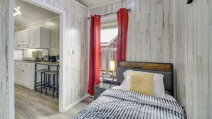 A bedroom with a bed, a nightstand, and a window with red curtains. There is a kitchen visible through an open door. - Cozy Picayune Rental Home ~ 11 Mi to Space Center! (Picayune)