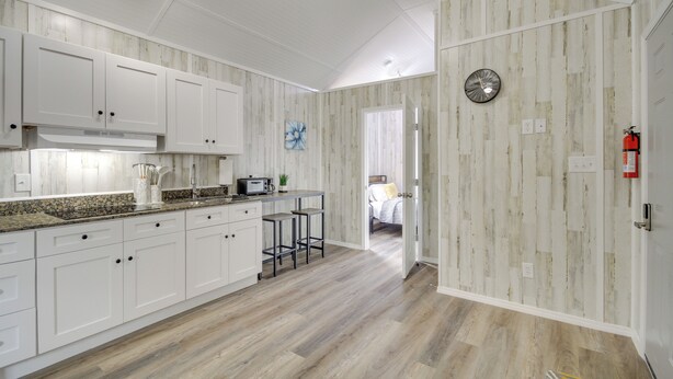 A kitchen with white cabinets, a granite countertop, and a microwave. There are two bar stools and a small table with a plant. The floor is wooden, and the walls have a wood panel design. - Cozy Picayune Rental Home ~ 11 Mi to Space Center! (Picayune)