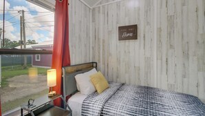 A bedroom with a bed, a nightstand, a lamp, a window with curtains, and a sign on the wall. - Pet-friendly Picayune Tiny Home Near Space Center! (Picayune)