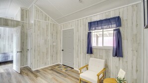 A room with wooden panel walls, a white door, a window with blue curtains, a white chair, and a small table with a plant. - Pet-friendly Picayune Tiny Home Near Space Center! (Picayune)