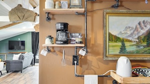 Une cuisine avec des étagères en bois où sont rangés les ustensiles de cuisine, une machine à café et une bouilloire. Un tableau représentant un paysage de montagne est accroché au mur.