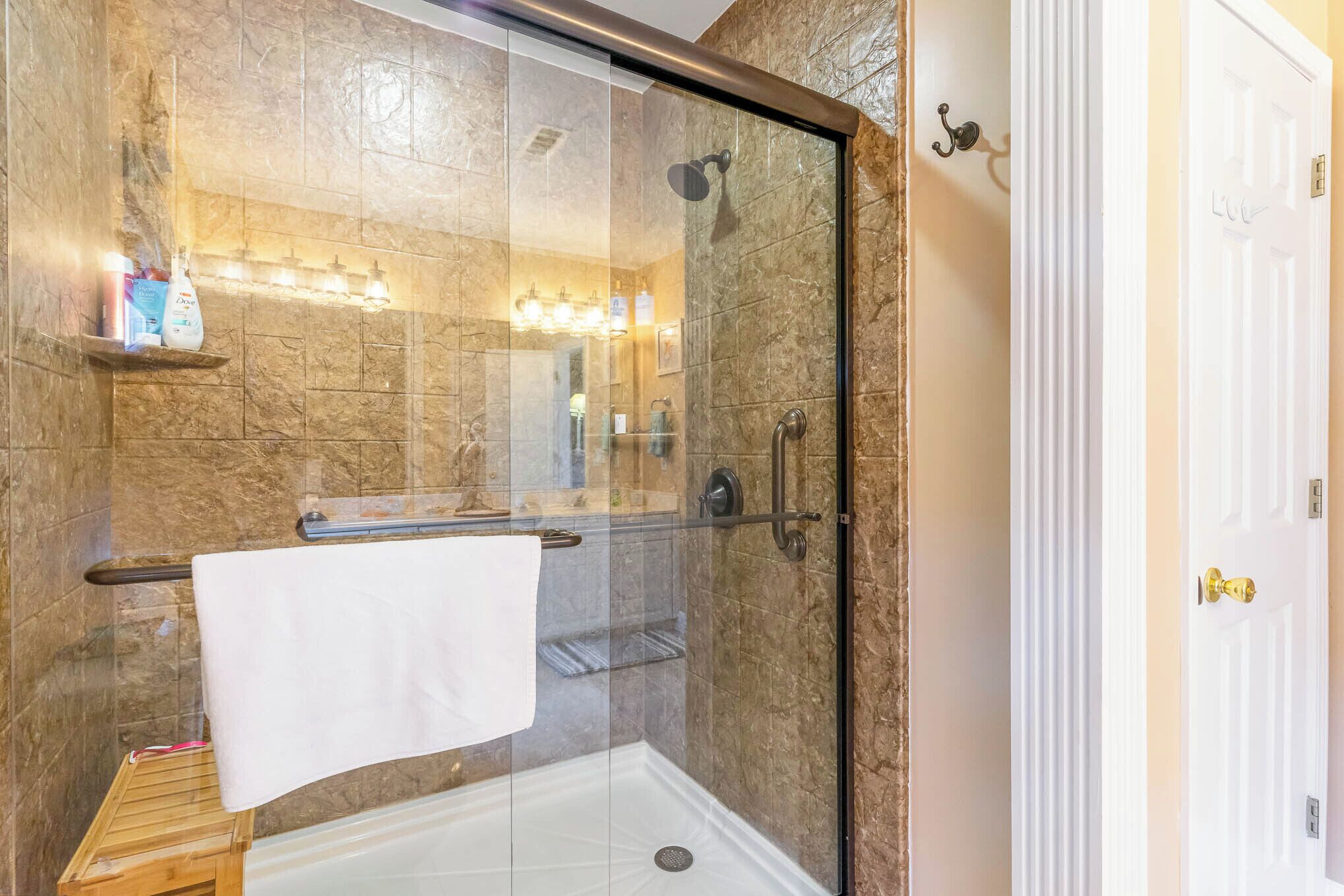 Une douche fermée par des parois de verre, avec une serviette blanche suspendue à un porte-serviettes, un tabouret en bois et une étagère contenant des articles de toilette.