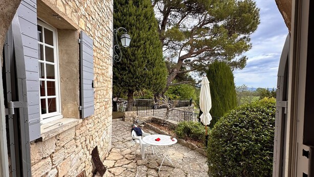 A stone pathway leading to a patio area with a table and chairs, a stone wall, and a view of trees and a distant landscape. - From the top of its terraces and garden, breathtaking views of the Luberon  (Murs)