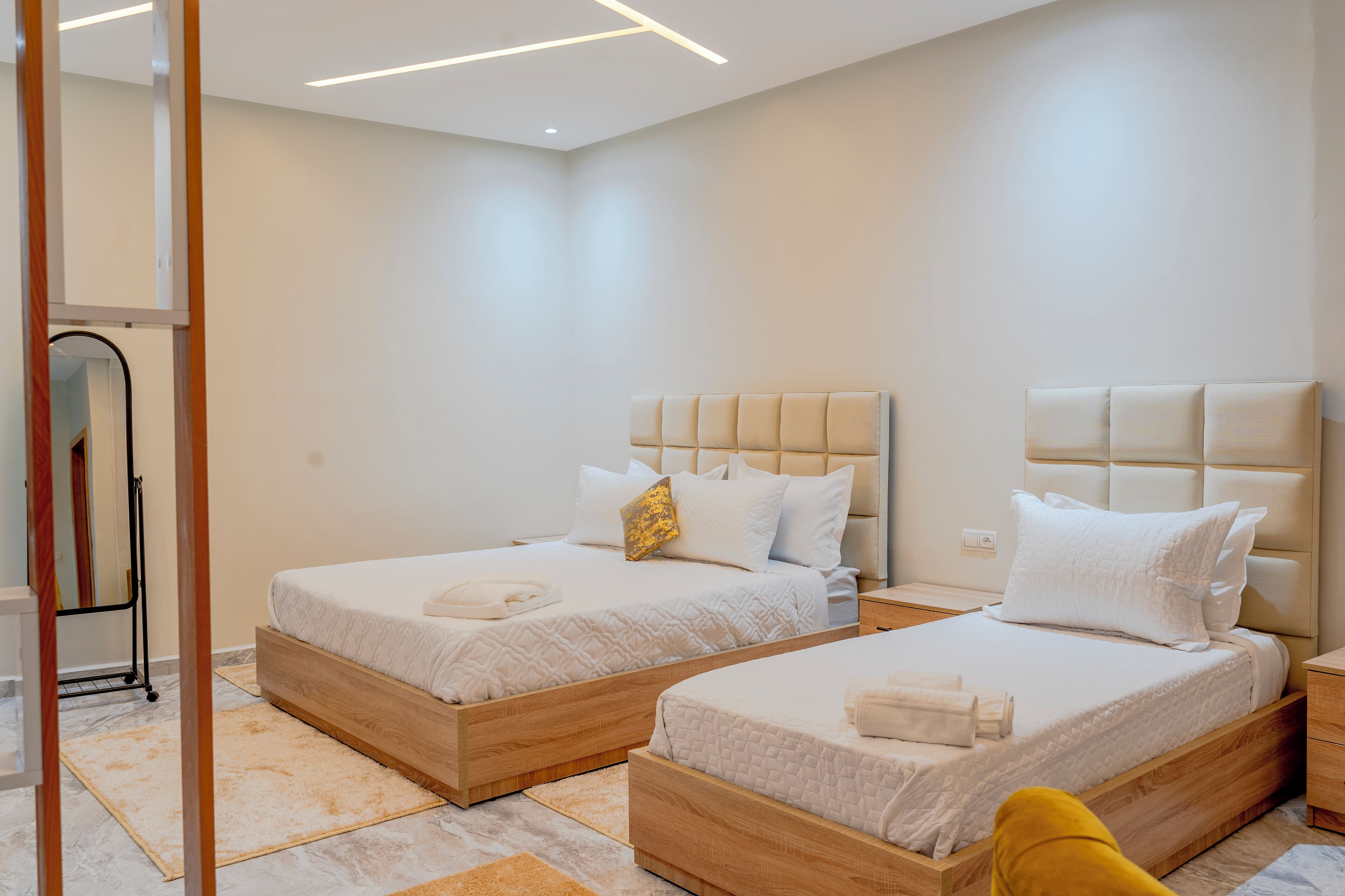 Habitación de hotel con dos camas, cada una con estructura de madera y ropa de cama blanca. Hay una mesita de noche pequeña con una lámpara y un enchufe en la pared.