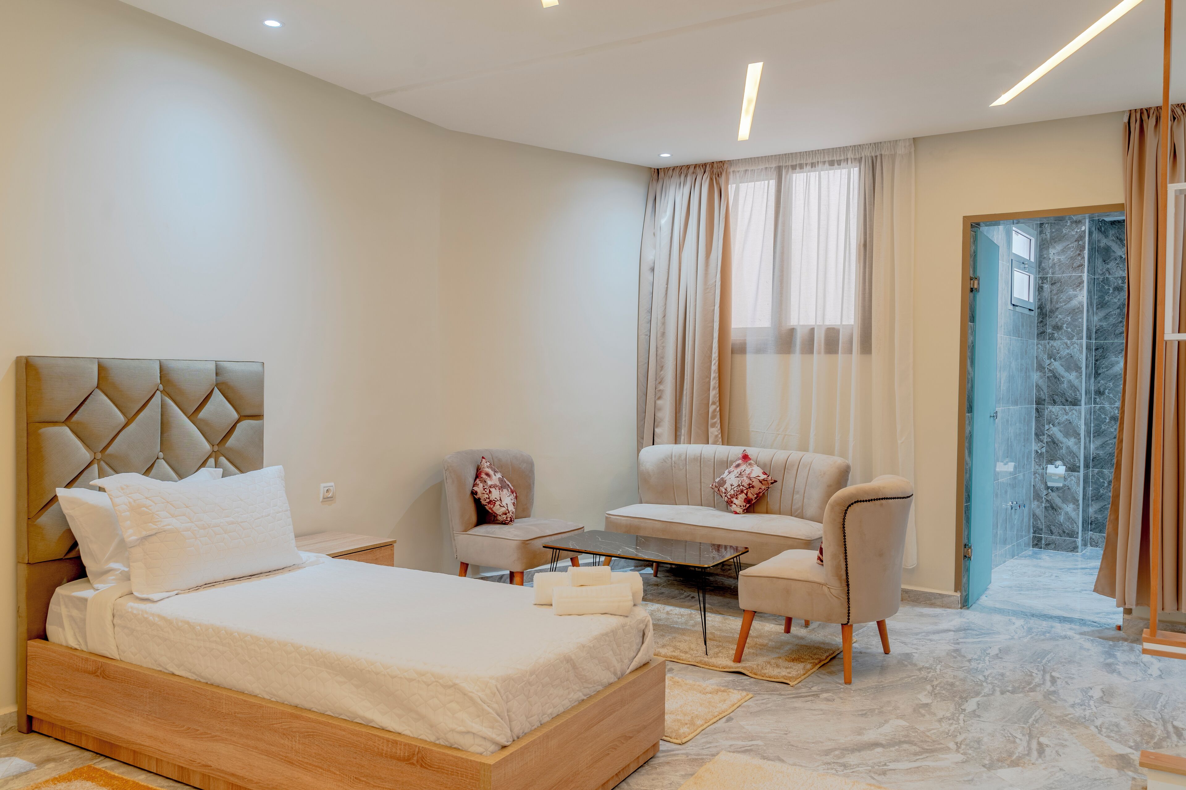 A modern hotel room with a large bed, two armchairs, a small table, and a glass-enclosed shower area.