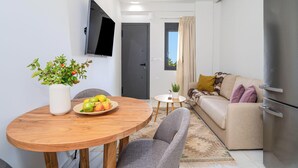 A modern living room with a sofa, a dining table with a fruit bowl, a TV mounted on the wall, and a refrigerator. - Inia Apartments (Malevizi)