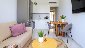 A compact living space with a sofa, a dining table with chairs, a kitchen area, and a TV mounted on the wall. - Inia Apartments (Malevizi)