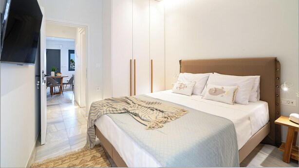 A bedroom with a bed, bedside table, and a TV mounted on the wall. - Inia Apartments (Malevizi)