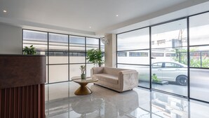 Un hall d’entrée moderne avec un canapé beige, une table basse en verre et un élément décoratif en bois. De grandes fenêtres offrent une vue sur la rue et sur un bâtiment portant l’inscription « Tribunal 1 ».