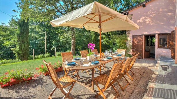 Un área de comedor al aire libre con una mesa dispuesta para cuatro personas, rodeada de sillas de madera, bajo un paraguas grande.