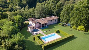 Una casa grande con piscina, rodeada de césped cuidado y árboles frondosos.