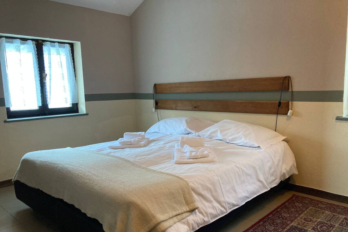 A neatly made bed with white linens and towels, a wooden headboard, a patterned rug, and a window with sheer curtains.