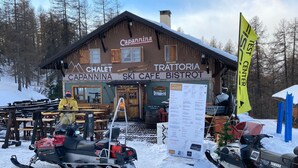 Un café de esquí con fachada de madera, paisajes nevados y motos de nieve estacionadas adelante.