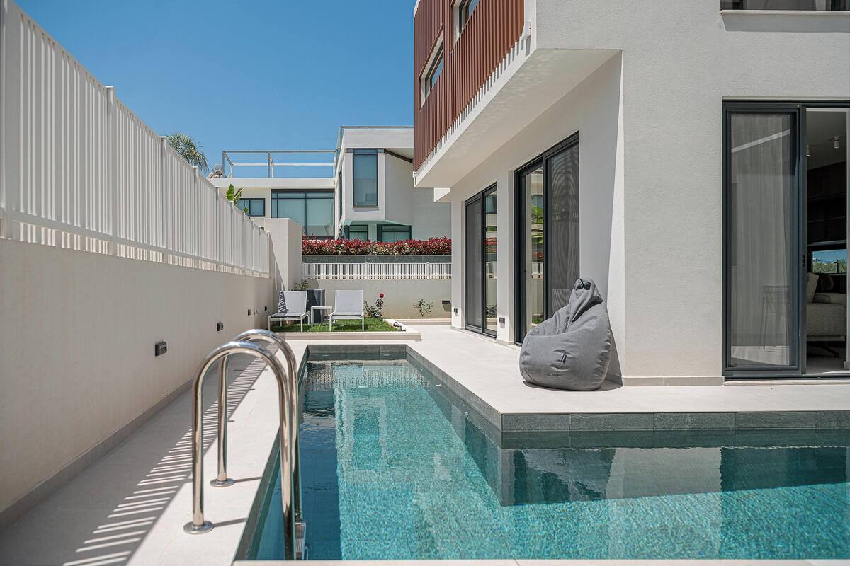 Uma área de piscina ao ar livre moderna, com piscina azul límpida, escada de metal e área de descanso com um bean bag cinza.