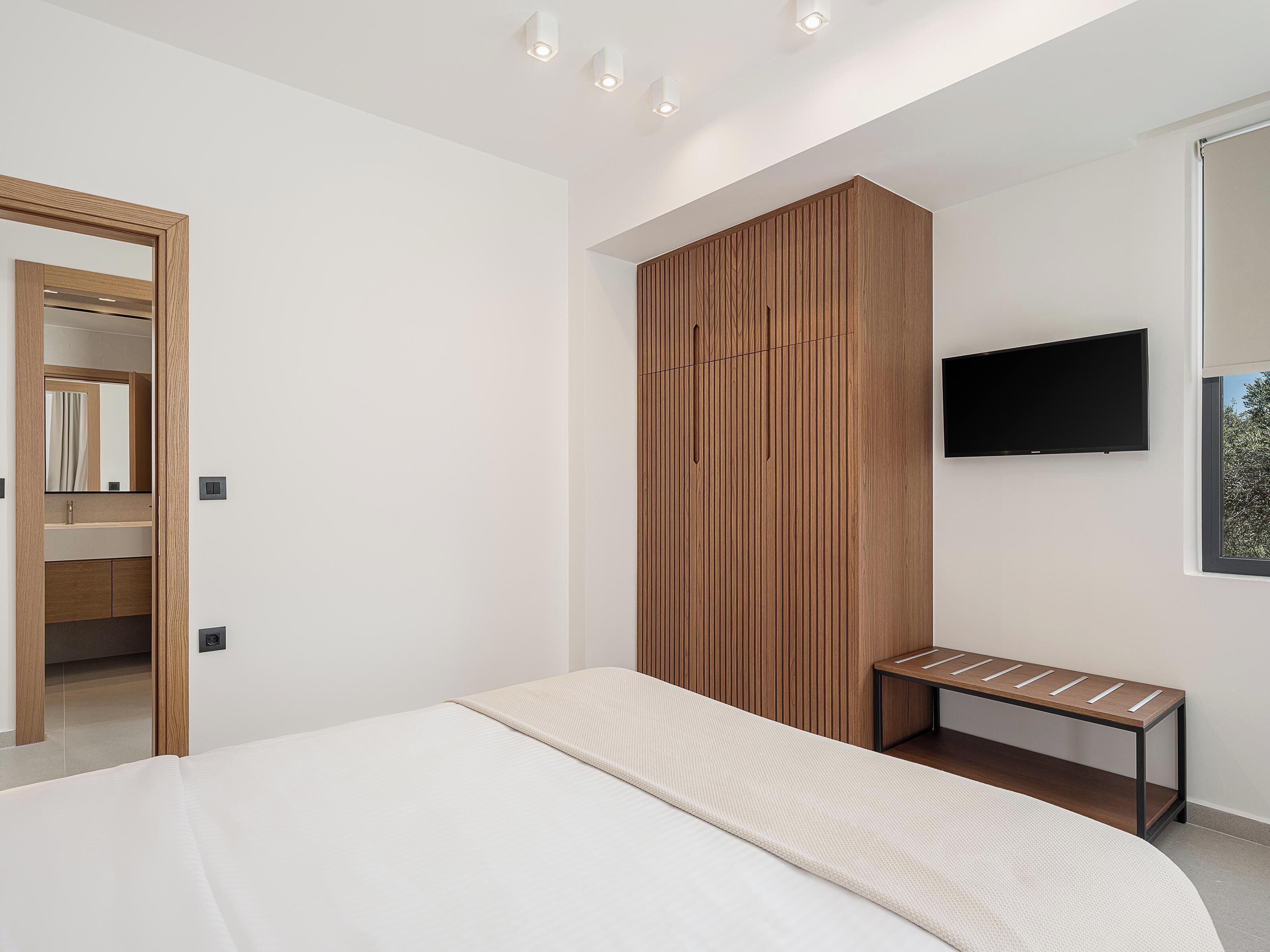 A modern bedroom with a bed, a wooden wardrobe, a bench, and a TV mounted on the wall.