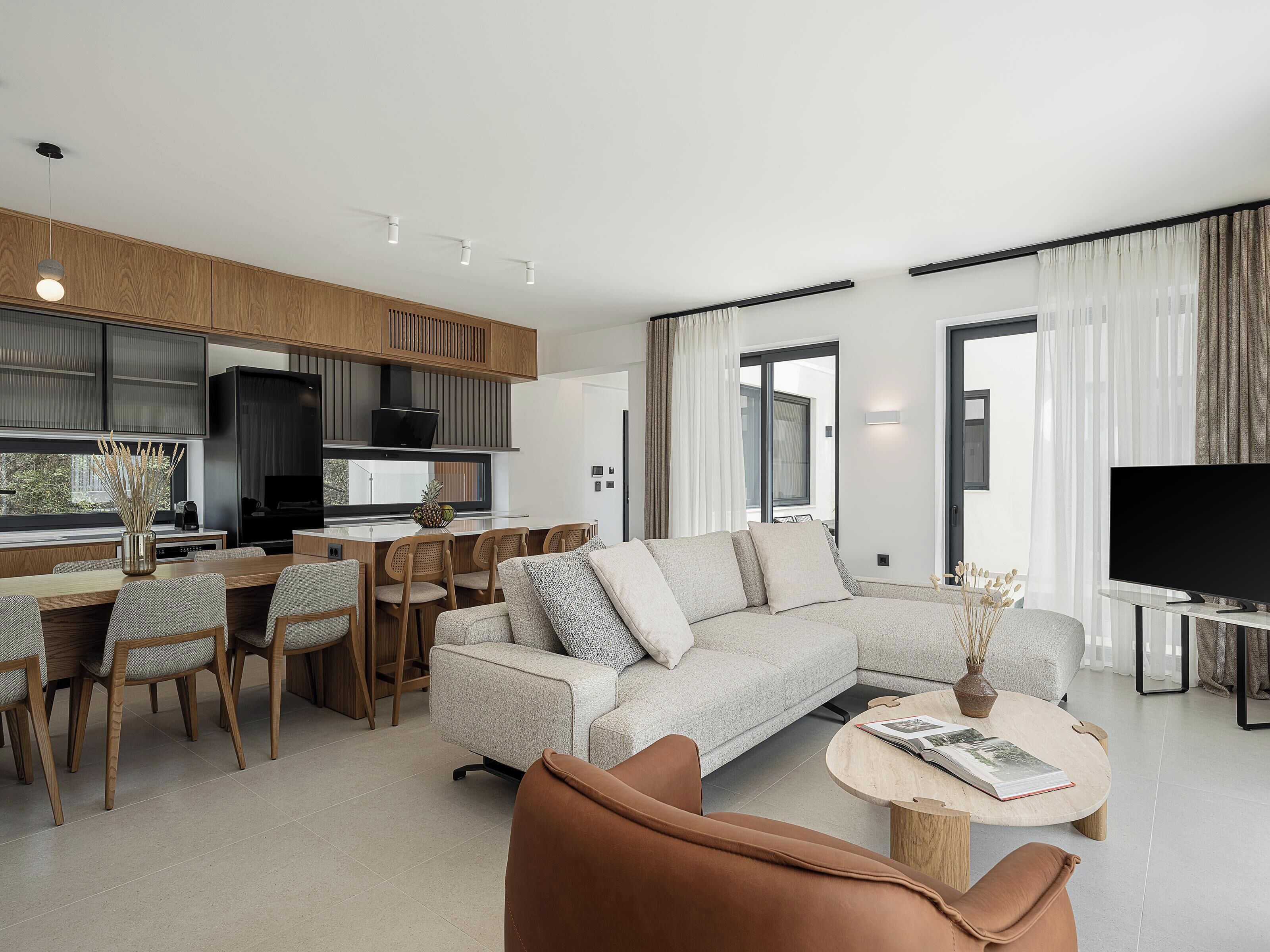 Ein modernes Wohnzimmer mit einer großen Eckcouch, einem Couchtisch und einem Fernseher. Es gibt einen Essbereich mit Stühlen und einem Tisch, sowie eine Küche mit Holzschränken und einem schwarzen Kühlschrank.