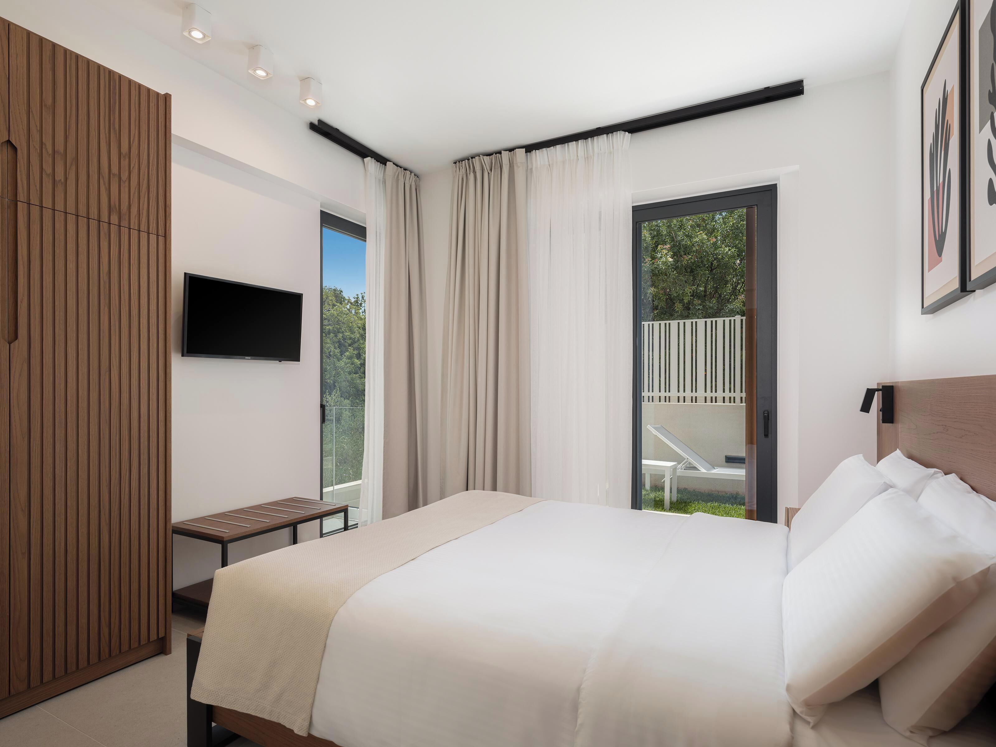 Une chambre moderne avec un grand lit, un téléviseur fixé au mur, une armoire en bois et un balcon donnant sur un paysage verdoyant.