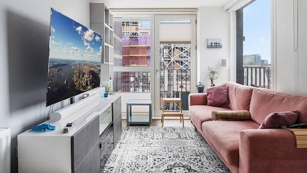 Un salon moderne doté d’un téléviseur à écran plat, d’un canapé rose, d’un tapis à motifs et offrant une vue sur le paysage urbain à travers une porte vitrée.