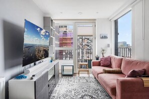 A modern living room with a flat-screen TV, a pink sofa, a patterned rug, and a view of a cityscape through a glass door.