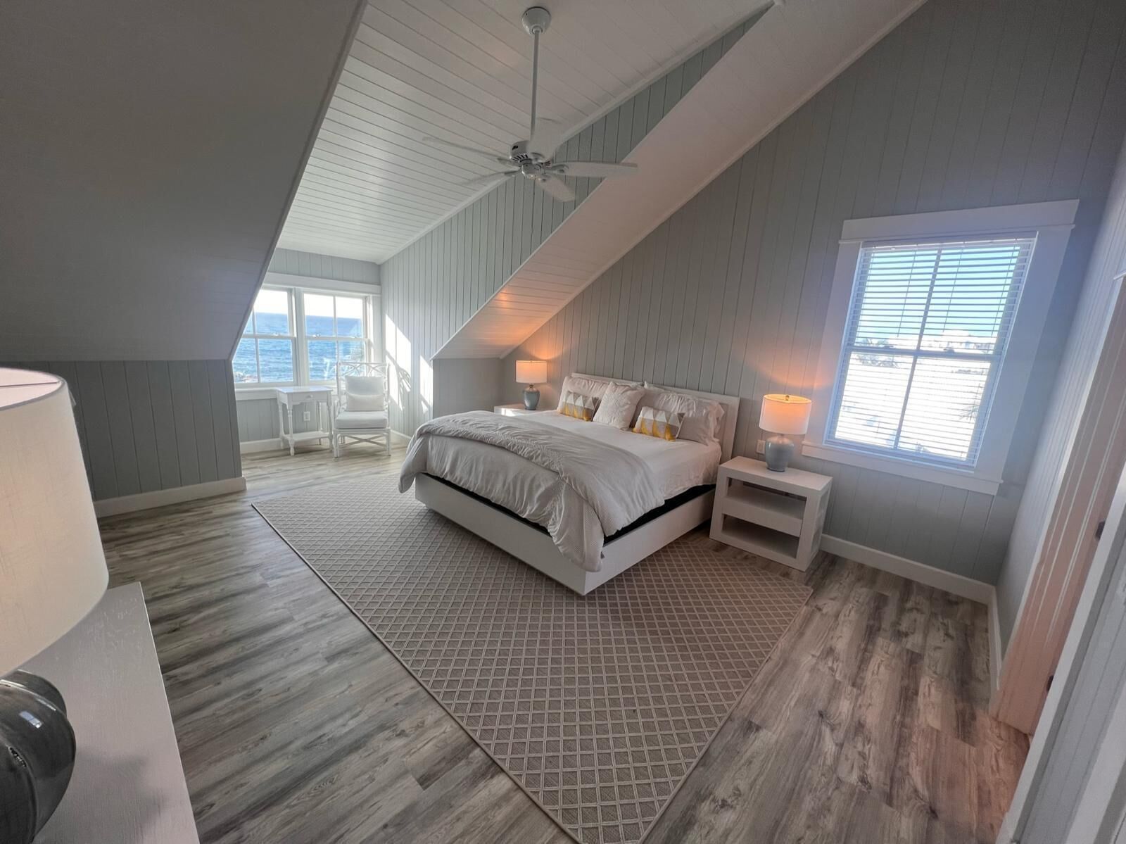 A bedroom with a large bed, a desk, and a chair. There is a window with blinds and a view of the outdoors.