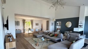 Amplia sala de estar con un sofá grande de color gris, una mesa de centro de vidrio y una mesita auxiliar de madera. En el fondo, se distingue un área de comedor con una mesa y sillas.