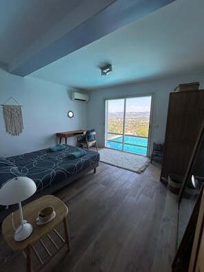 Una camera da letto con un letto, una scrivania, una sedia, un tavolino con una lampada e un armadio. C'è un'ampia finestra che offre vista su una città e montagne.