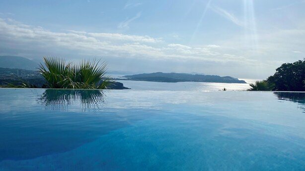 Piscina infinita azul cristalina con vistas al mar y montañas lejanas.