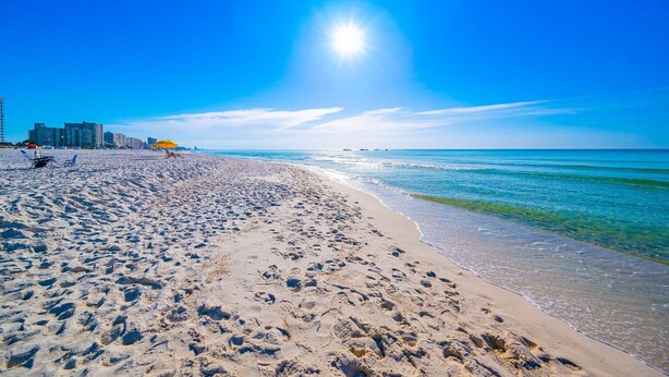 Une plage parsemée d’empreintes, un ciel dégagé et des bâtiments en arrière-plan.