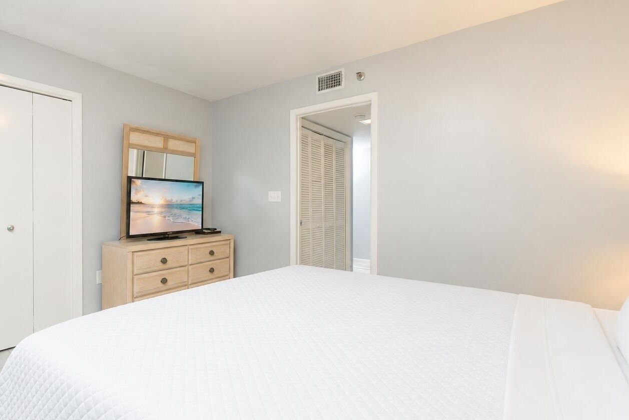 Un dormitorio con una cama, un aparador, un televisor y una puerta que da a otra habitación.