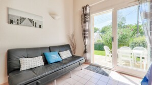 A modern living room with a black leather sofa, patterned cushions, a wall-mounted light, and a view of an outdoor patio with white furniture. - 350 m from the beach – House for 5 – Garden & BBQ (Sarzeau)