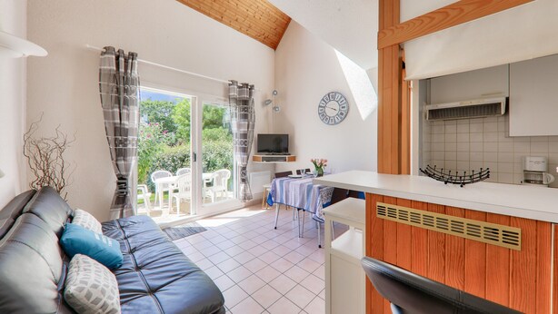 A modern living room with a sofa, a TV, and a kitchen area with a clock and a window. - 350 m from the beach – House for 5 – Garden & BBQ (Sarzeau)