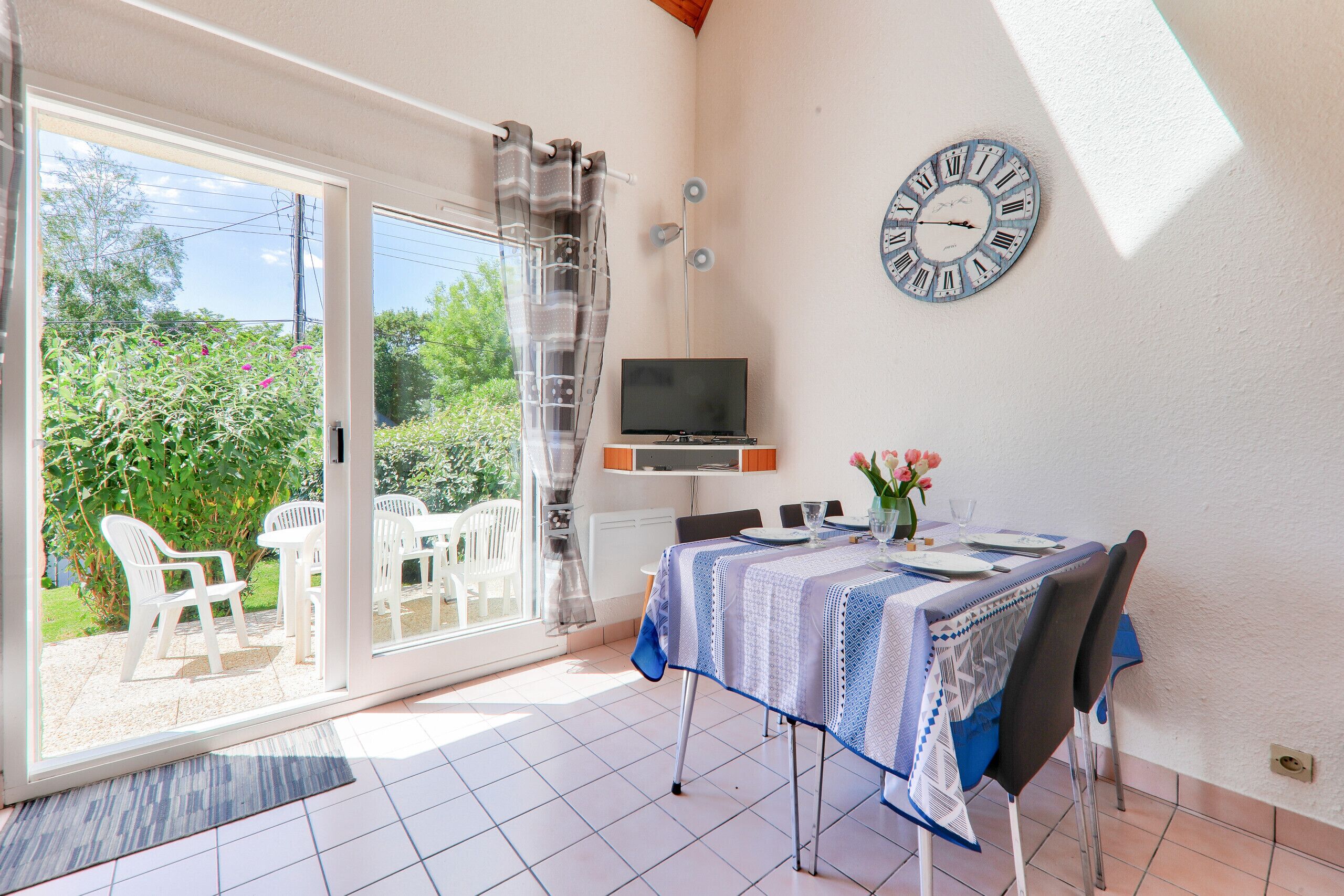 Un coin repas avec une table dressée pour deux personnes, un téléviseur posé sur un meuble et une horloge au mur.