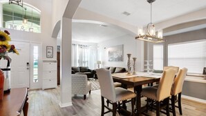 A spacious dining area with a wooden table and chairs, a chandelier, and a view of a living room with a sofa and a dining table.