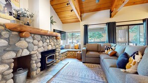 A cozy living room with a stone fireplace, a grey sofa, and a large window with curtains. - Bigwood #45 2 Bedroom Condo by RedAwning (Mammoth Lakes)