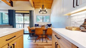 A modern kitchen with wooden cabinets, a dining table with chairs, a chandelier, and a large window with curtains. - Bigwood #45 2 Bedroom Condo by RedAwning (Mammoth Lakes)