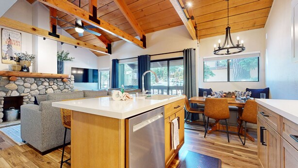 A modern kitchen with a central island, stone fireplace, wooden ceiling beams, and large windows offering views of the outdoors. - Bigwood #45 2 Bedroom Condo by RedAwning (Mammoth Lakes)