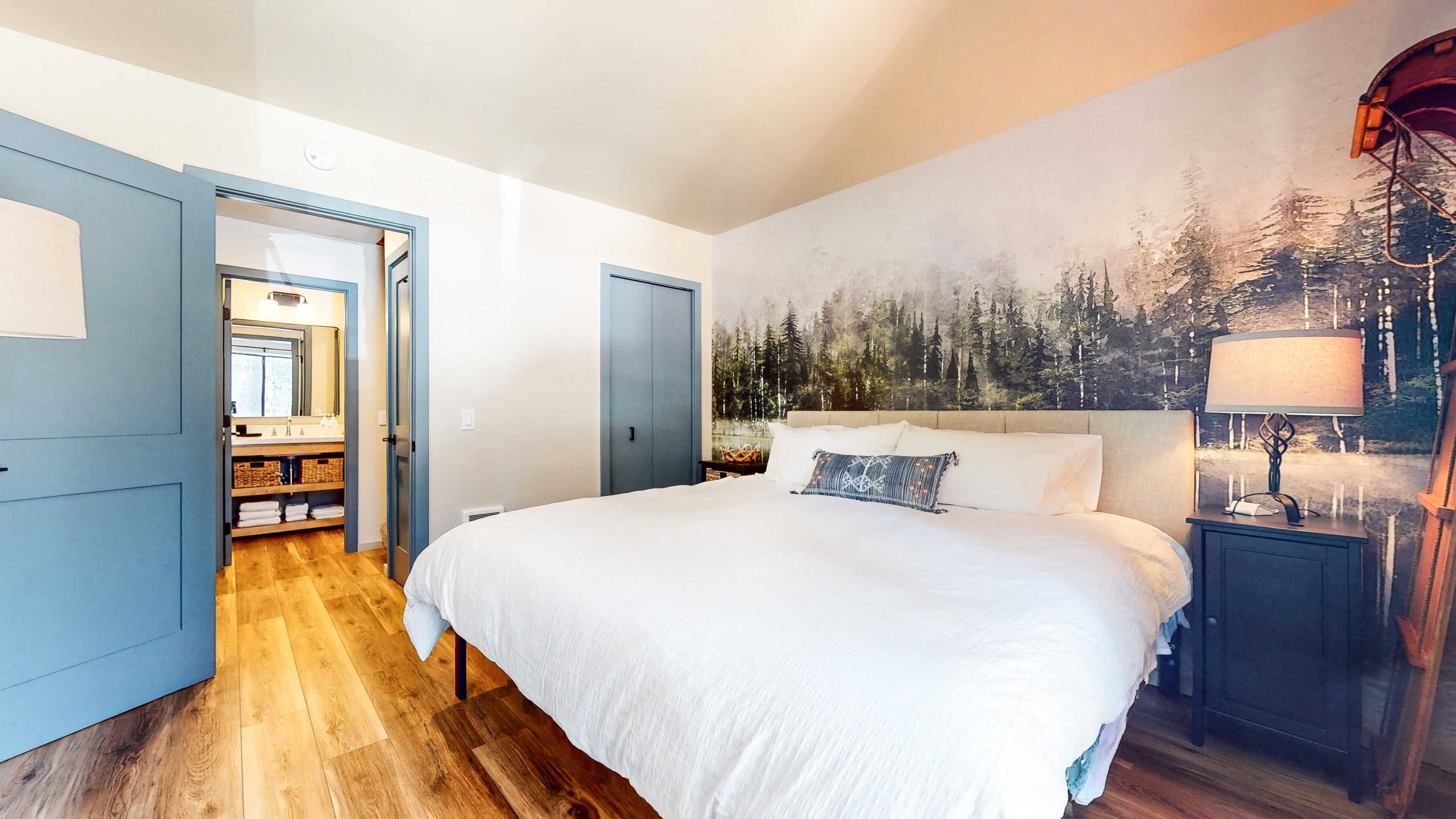 Un dormitorio con una cama grande, una mesita de noche, una lámpara y un mural de un bosque en la pared.