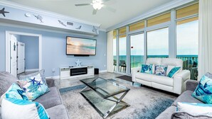 A living room with a TV, a glass coffee table, a white sofa with blue cushions, and a balcony with a view of the ocean.