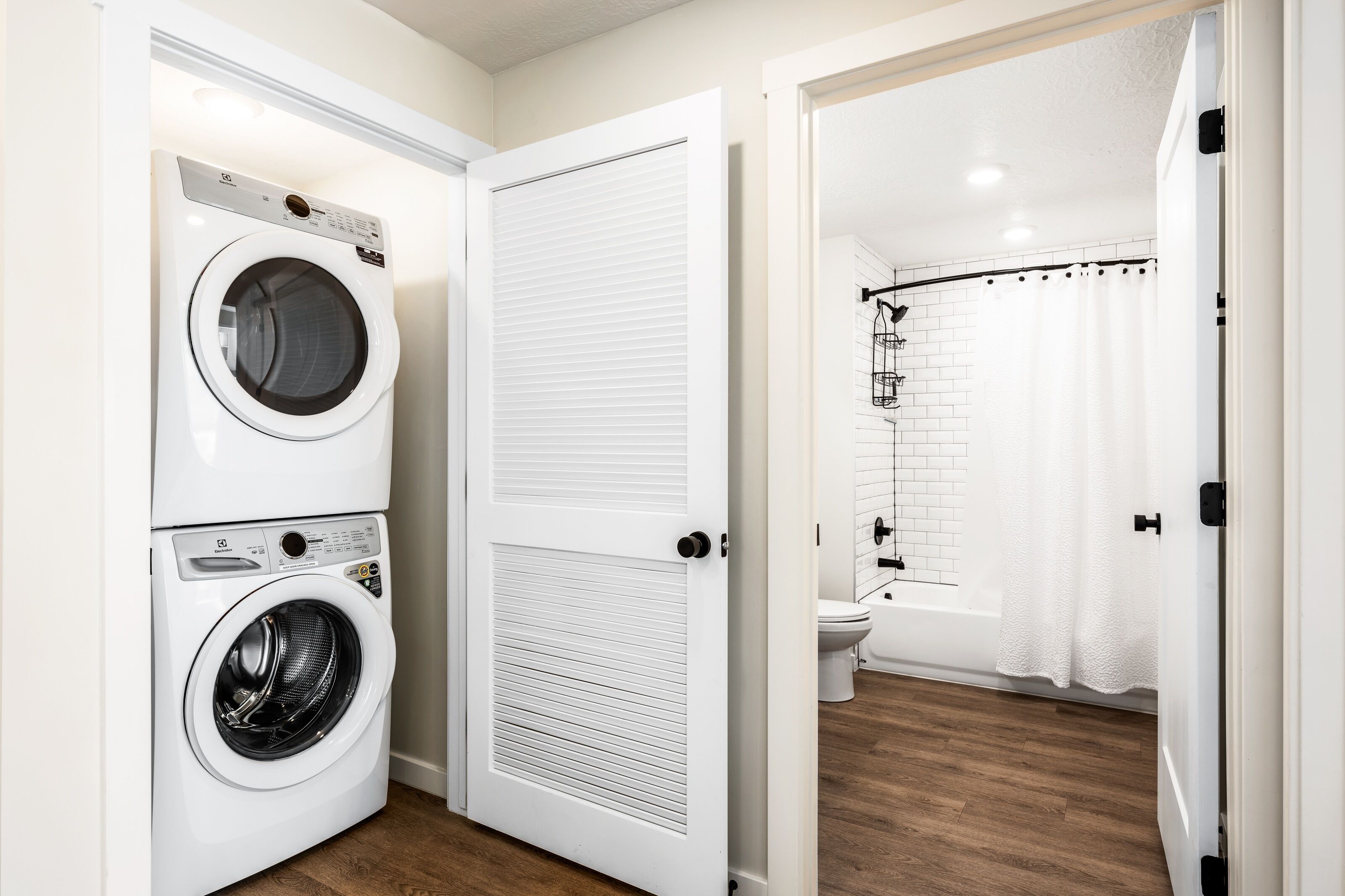 A laundry room with stacked washers and dryers, a closed door, and a view into a bathroom with a shower and toilet.