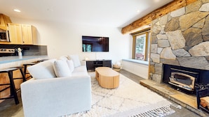 A modern living room with a stone fireplace, a sofa, a coffee table, and a kitchen area with a microwave. - Lakeview Villas #09 3 Bedroom Condo by RedAwning (Mammoth Lakes)