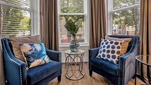 A room with two blue armchairs, a glass table with a vase, and a view through large windows with blinds.